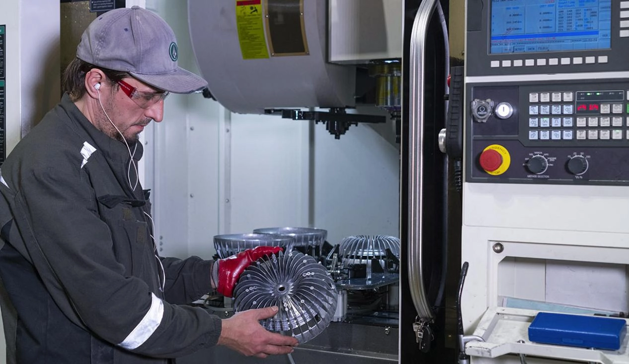 Factory worker (wearing cap, safety glasses, and work jacket) inspecting a metal fan component